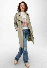 Hellgrauer Trenchcoat über einem gepunkteten pastellfarbenen Pullover, kombiniert mit verwaschenen Jeans und beigen Clogs, auf einem neutralen Hintergrund.