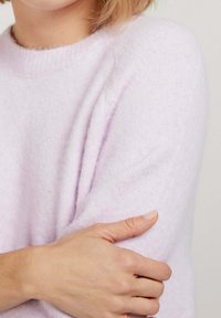 Pull léger violet, doux, avec un col rond, finition texturée et épaules tombantes. Main reposant sur le bras opposé.