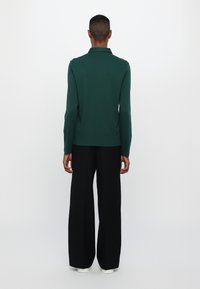 Mørk grønn langermet polo skjorte laget av myk stoff, kombinert med svarte vide bukser og hvite sneakers. Rent, minimalistisk design.
