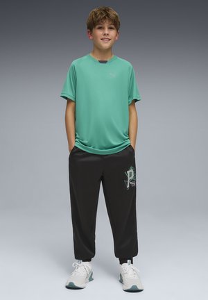 Niño de pie con las manos en los bolsillos, vistiendo una camiseta verde de Puma, pantalones negros con una gráfica de la letra P y zapatillas blancas, sonriendo a la cámara.