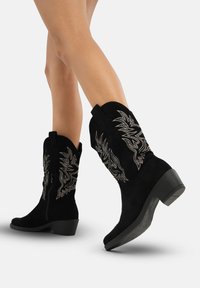 Las botas de vaquero de ante negro presentan patrones bordados en plata, un borde superior curvado y un tacón bajo en bloque para mayor estabilidad y comodidad.