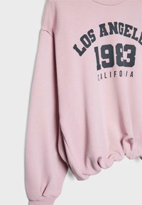 Svetloružový sveter s okrúhlym výstrihom, spustenými ramennými švami, elastickým lemom a výrazným čiernym dizajnom textu "LOS ANGELES 1983 CALIFORNIA".