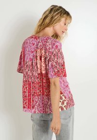 Blouse multicolore avec des motifs paisley et floraux en rouge, rose et crème ; manches courtes et coupe décontractée avec texture froncée.