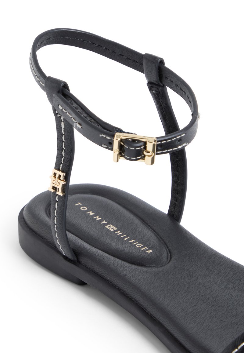 Sandalia plana negra con correa en el tobillo, hebilla dorada, costuras blancas y la marca "Tommy Hilfiger" en la suela acolchada.