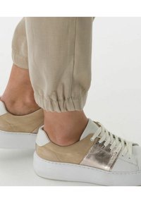 Sneakery mają białą gumową podeszwę, beżową cholewkę z zamszu i skóry, metaliczne akcenty oraz teksturalne detale sznurowadeł; elastyczne spodnie z mankietami w jasnym beżu.