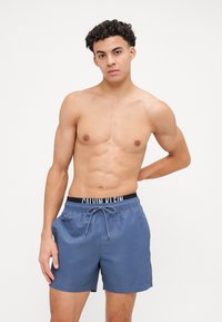 Zilas peldkostīms, kas izgatavots no viegla materiāla ar elastīgu jostu, kurai ir melns Calvin Klein zīmols un regulējams aukliņš.