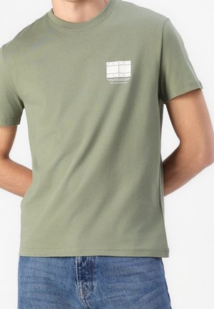Person trägt ein olivgrünes Tommy Jeans T-Shirt mit kleinem weißem Logo auf der Brust und blaue Denim-Jeans, Hände hinter dem Rücken.