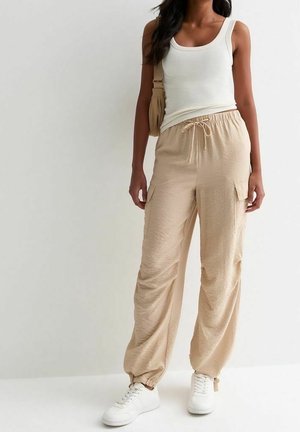 Frau trägt beige Cargohose, weißes Tanktop, weiße Sneakers und trägt eine beige Umhängetasche, steht vor einem einheitlichen Hintergrund.