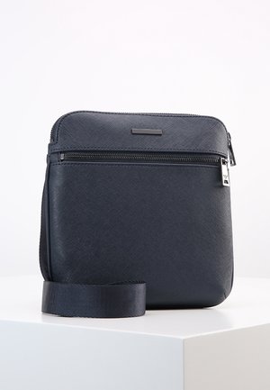 Compact marineblauwe crossbodytas met een gestructureerd oppervlak, een voorvak met rits en een verstelbare band, geplaatst op een wit voetstuk.