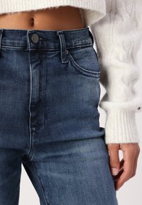 Høytlivs blå denim jeans har knappelukking, fem lommer, og lett falming. Kombinert med en kremfarget strikkegenser.