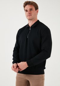 Fekete cipzáros pulóver hosszú ujjal, gallérral, bordázott mandzsettával és aljjal. Simább anyagtextúra, testre simuló fazon, világosbézs nadrág felett viselve.