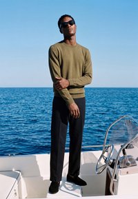 Homme portant des lunettes de soleil, un pull olive et un pantalon noir, debout sur un bateau avec l'océan et un ciel clair en arrière-plan.