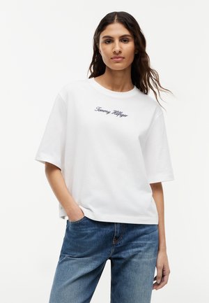 Νεαρή γυναίκα με μακριά σκούρα μαλλιά που φοράει μια λευκή μπλούζα Tommy Hilfiger και μπλε τζιν, στέκεται με το ένα χέρι στην τσέπη.