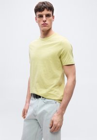 Νεαρός άνδρας που φοράει ανοιχτόκίτρινη κοντομάνικη μπλούζα και ανοιχτό μπλε τζιν με μαύρη ζώνη, στέκεται μπροστά σε απλό λευκό φόντο.