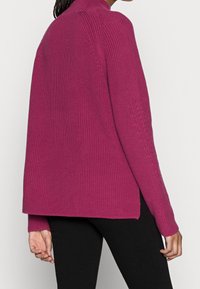 Magenta gebreide sweater met een hoge kraag, ribbelstructuur en zijsplitsen. Beschikt over lange mouwen met kabelbrei-accenten op de schouders.