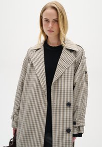 Trench coat a quadri beige e blu navy con ampio colletto, bottoni doppi e tasche laterali, indossato sopra un maglione nero con texture.