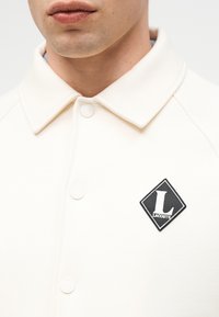 Homme portant une chemise écrue à col boutonné avec un écusson logo Lacoste noir en forme de losange sur la poitrine gauche.