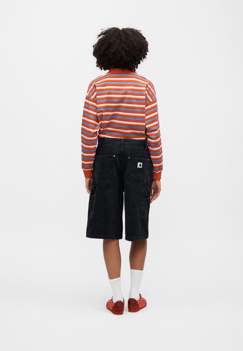 Person steht mit dem Rücken zum Betrachter, trägt ein orange gestreiftes Langarmshirt, schwarze Shorts, weiße Socken und rote Schuhe.