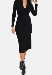 Svart rillad omslag klänning med långa ärmar, v-ringning och knytning i midjan, som når till mitten av vadbenet, tillsammans med svarta ankelstövlar.