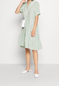 Abito floreale verde con maniche corte, design a strati e abbottonatura frontale. Indossato con mule bianche e una piccola borsa a tracolla.