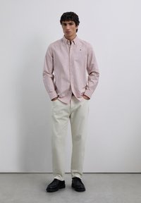 Rotes und weiß gestreiftes Hemd mit langen Ärmeln, kombiniert mit cremefarbenen Hosen und schwarzen Schuhen, stehend vor einem neutralen Hintergrund.