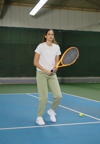 Sieviete tenisa spēlētāja, ģērbusies baltā T-kreklā un zaļās sporta biksēs, turot apelsīnu tenisa raketi, uz zila laukuma ar tuvumā esošu tenisa bumbu.