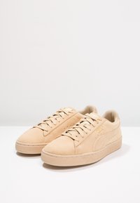 Un paio di sneaker in suede beige con lacci, suola testurizzata e branding sottile su una superficie bianca uniforme.