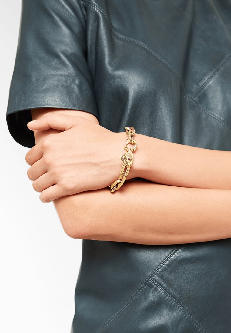 Le bracelet en chaîne dorée présente des maillons épais et un pendentif en forme de diamant. L'arrière-plan comprend un haut en cuir noir avec une texture lisse.