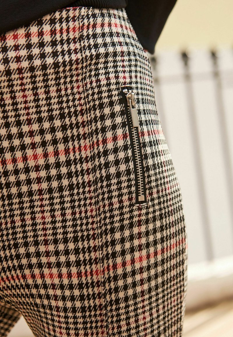 Gros plan d'un pantalon à motif pied-de-poule avec des lignes noires, blanches et rouges et une fermeture éclair noire verticale au niveau de la hanche.