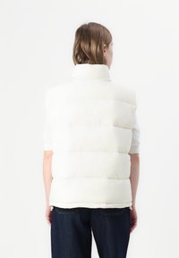 Witte puffer gilet met een hoge kraag, uitgerust met gequiltte secties en een gladde textuur. Draagt over een shirt met korte mouwen, gecombineerd met donkere jeans.
