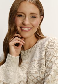 Krémový sveter s diamantovým vzorom v béžovej farbe, s rebrovaným golierom a manžetami. Modelka má dlhé vlasy a usmieva sa.