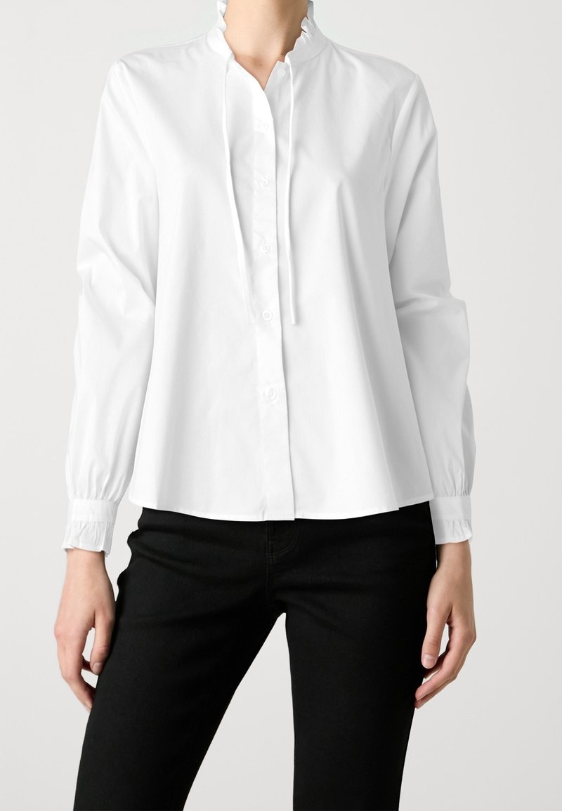 Femme portant un chemisier blanc à manches longues avec boutonnière à l'avant et col à volants, associé à un pantalon noir, debout devant un fond uni.