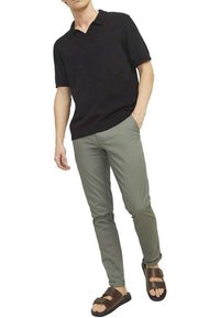 Polo shirt noir à manches courtes, pantalon slim-fit vert clair, et sandales brunes à deux brides, portés par une figure masculine en position debout.