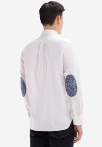 Chemise blanche à manches longues avec des coudières en tissu floral bleu à motifs, un empiècement au dos, fabriquée dans un tissu tissé lisse.