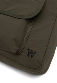 Sac rembourré vert olive avec un rabat, des bords surpiqués et un logo "W" brodé en noir. Tissu durable avec une texture lisse.