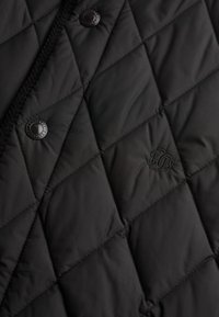 Chaqueta acolchada negra con un patrón de diamantes, botones a presión y un sutil logotipo bordado. El material parece suave y ligero.