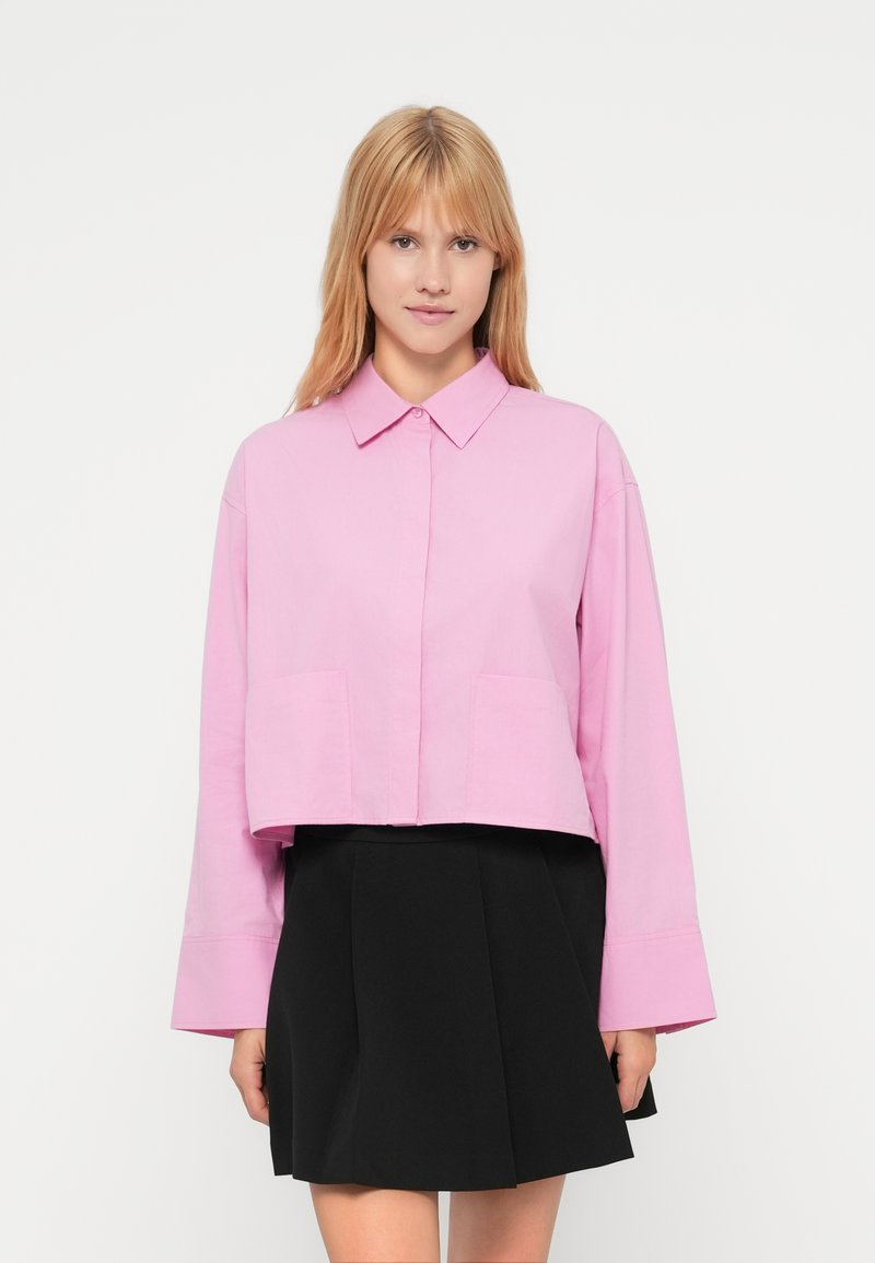 Ροζ cropped πουκάμισο με μακριά μανίκια, γιακά με κουμπιά και δύο τσέπες εμπρός, σε συνδυασμό με μαύρη φούστα. Υφασμα με υφή.