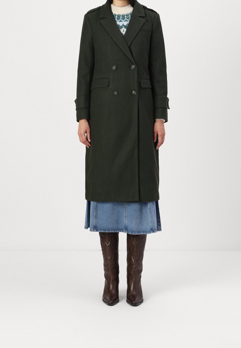 Femme portant un manteau vert foncé à double boutonnage par-dessus un pull à motifs, une jupe en jean et des bottes en cuir marron montant jusqu'aux genoux.