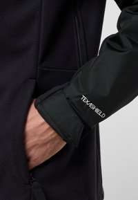 Giacca in pile nera con tessuto testurizzato nero a contrasto sui polsini. Presenta una tasca con zip e il marchio "TEXASHIELD" sulla manica.