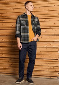 Camicia di flanella a quadri navy con accenti grigi e gialli, indossata sopra una t-shirt senape, abbinata a jeans scuri e stivali marroni. Sfondo in legno.