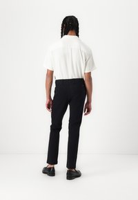 Camisa blanca de manga corta y textura, combinada con pantalones negros a medida y zapatos negros. La camisa tiene un cuello con solapa y cierre posterior con botones.