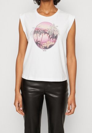 Witte mouwloze top met een grafisch ontwerp van een zonsondergang boven palmbomen met de tekst "Los Angeles" en sprankelende accenten. Gekoeld met zwarte broek.
