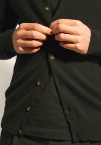 Svart kofta med texturerad yta, med guldknappar. Tyget hänger löst runt midjan, med en subtil fålldetalj.