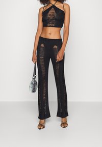 Kvinna i svart stickad haltertop och matchande stickade byxor med vertikala utklippta mönster, som håller en liten svart-vit mönstrad väska.