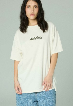 Giovane donna con lunghi capelli scuri che indossa una maglietta bianca oversize con la scritta "GRMY" e jeans blu con fantasia, su uno sfondo semplice.