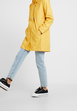 Person iført en gul regnfrakke, lyseblå jeans og sorte sneakers, der træder frem på en ensartet lys baggrund.
