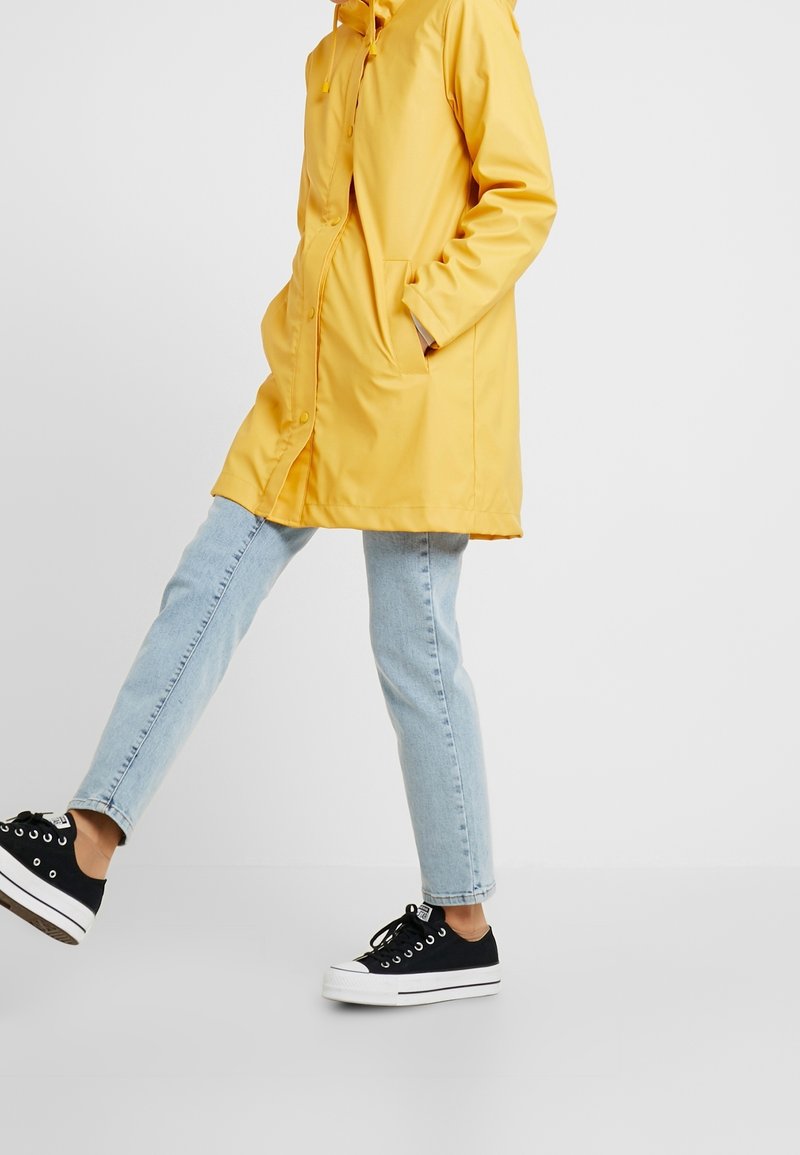 Personne portant un imperméable jaune, un jean bleu clair et des baskets noires marchant en avant sur un fond clair et uni.