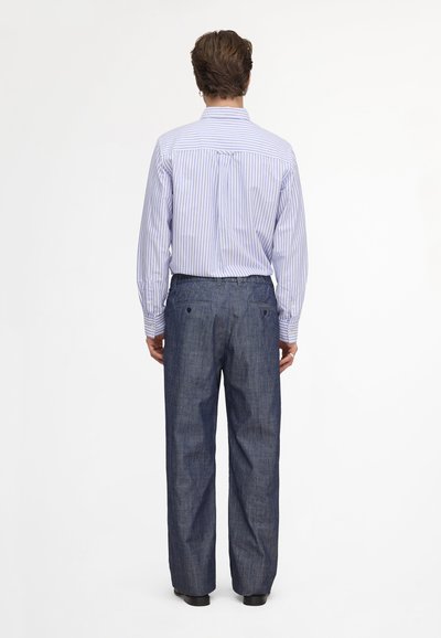 Homme debout de dos, portant une chemise à manches longues rayée bleu clair rentrée dans un pantalon ample bleu foncé avec des chaussures noires.
