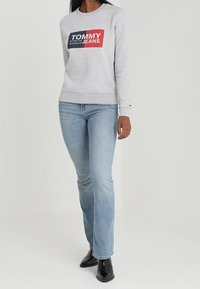 Grå sweatshirt med en stor röd och marinblå logotyp med texten "TOMMY JEANS", matchad med ljusblå jeans med raka ben och svarta stövlar.