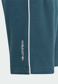 Tmavě modrý textil s bílou vertikální výpustkou a bílou výšivkou loga Adidas blízko lemu oděvu.